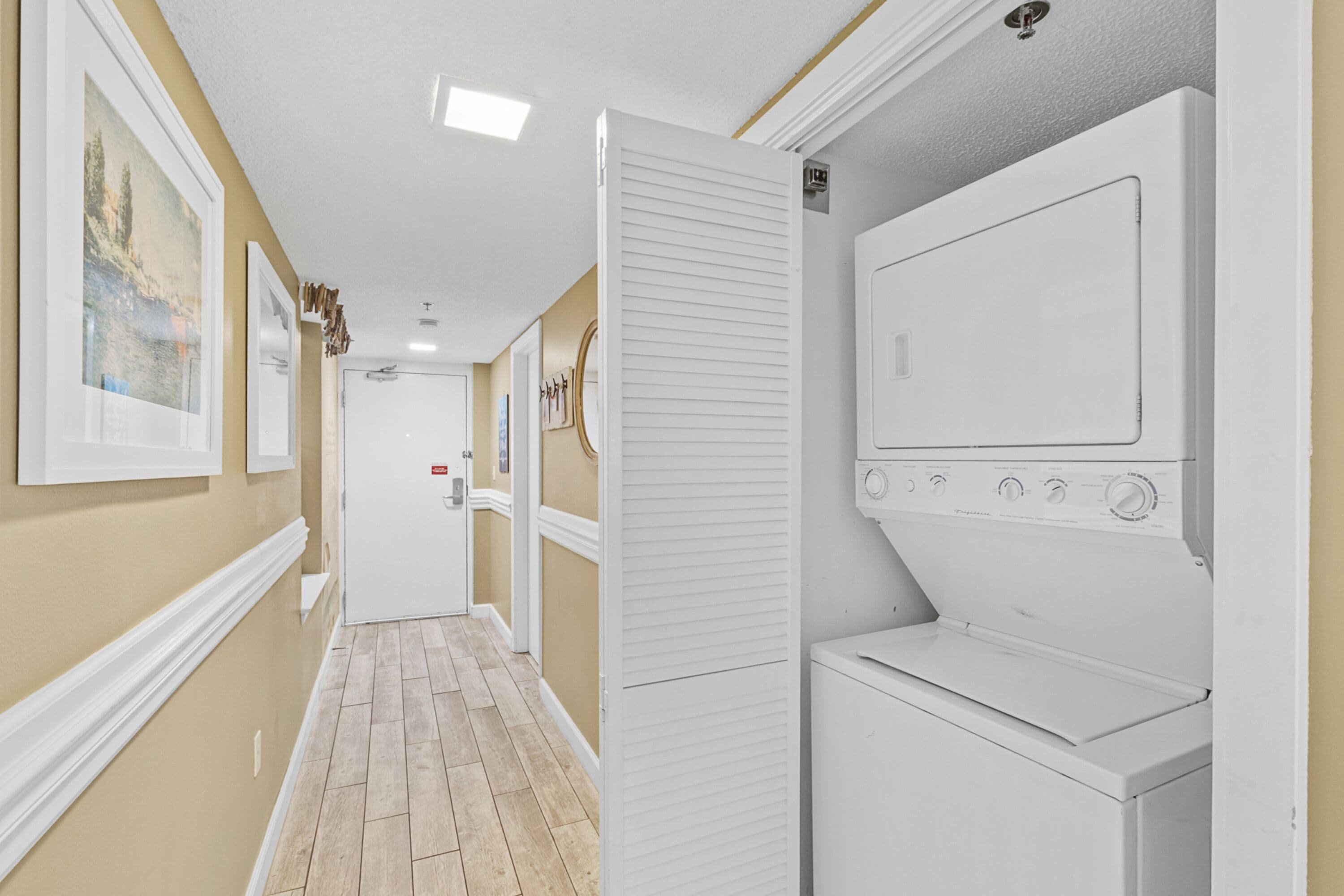 A laundry room with a front-loading washer and dryer, a door with a light switch, and a hallway with wooden flooring and framed pictures on the wall.