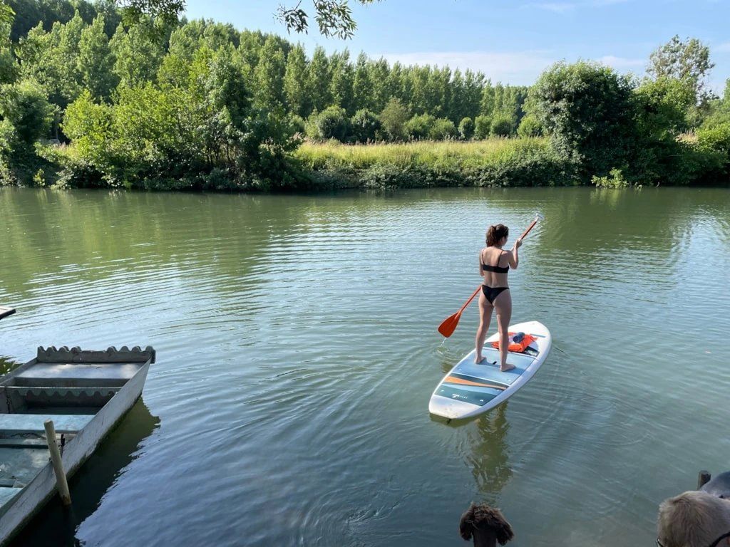 Une personne fait de la planche à pagaie sur une rivière paisible, avec un quai sur la gauche et une végétation luxuriante en arrière-plan.