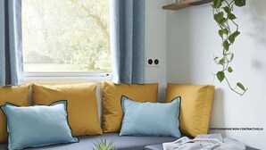 A cozy living room with a grey sofa, mustard and light blue cushions, a small table with a plant, books, and a kettle.