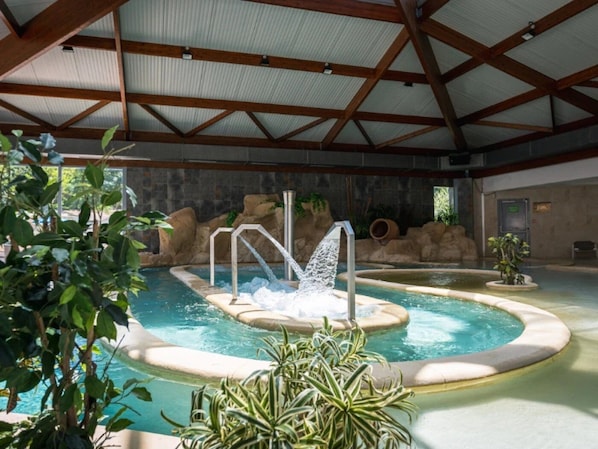 An indoor swimming pool with a hot tub, surrounded by plants and a stone wall.