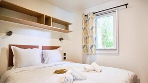 A hotel room with a bed, a window with curtains, a wooden shelf above the bed, and a small table with a book and towels. - ZEcamping 4* piscine chiens acceptés locatif clim 22m2 4 pers (Orgon)
