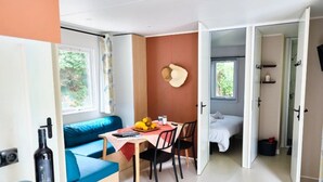 A room with a blue sofa, a dining table with chairs, a bed, and a window with a view of greenery. - ZEcamping 4* piscine chiens acceptés locatif clim 22m2 4 pers (Orgon)