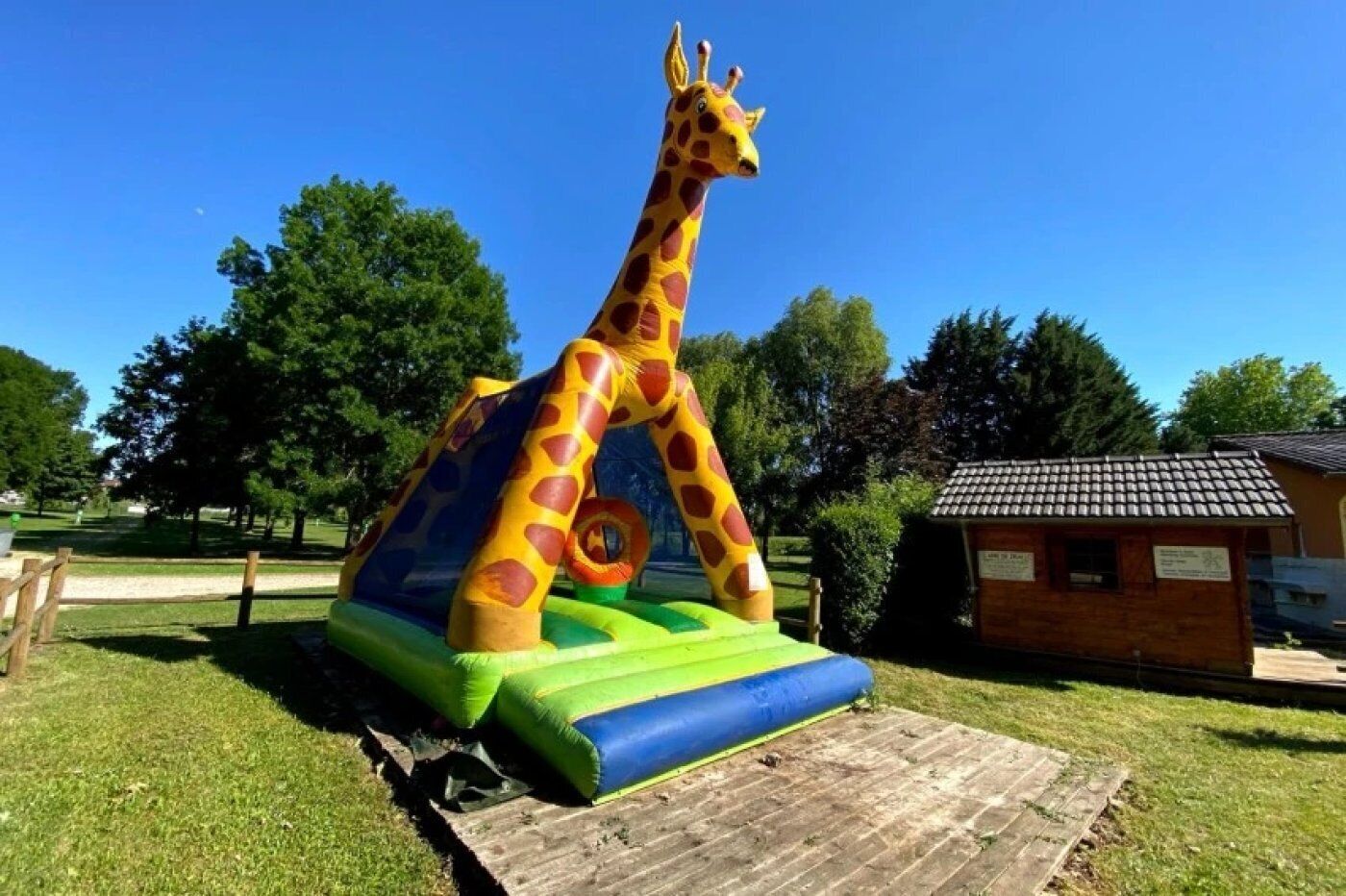 Une structure gonflable en forme de girafe, installée dans un parc avec des arbres et un petit bâtiment en bois en arrière-plan.