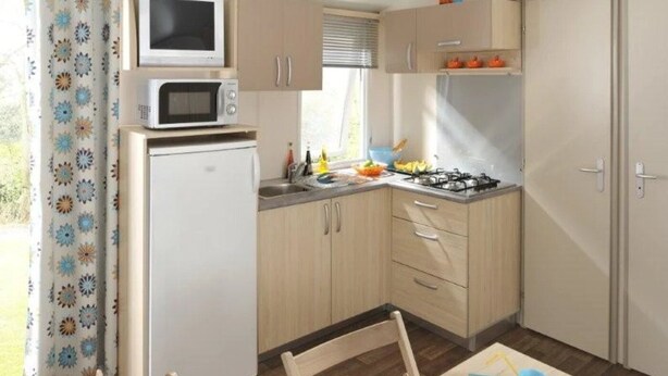 Une cuisine compacte équipée d’un four à micro-ondes, d’une cuisinière et d’un réfrigérateur. On y trouve une fenêtre avec des rideaux, une table avec des tasses et une chaise.