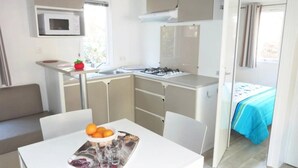 A compact kitchen with a microwave, sink, and gas stove. A dining table with a fruit bowl and glasses is in the foreground. - ZEcamping 3* 200m plage chiens acceptés locatif TV 36m2 6 pers (Hyères)