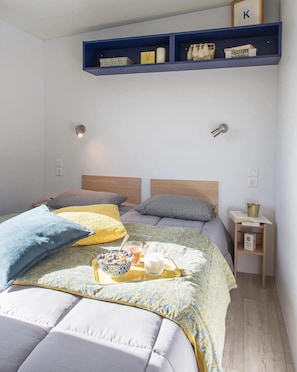 A bedroom with two beds, a bedside table with a book and a cup, and a shelf above the beds.