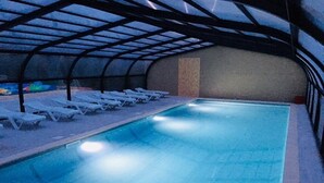 An indoor swimming pool with a clear blue water surface, surrounded by white lounge chairs and a covered roof structure. - ZEcamping 3* avec piscine chauffée club enfant animations pet friendly (Erquy)