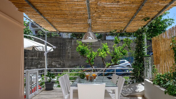 Eine Terrasse im Freien mit Tisch und Stühlen, einer hängenden Leuchte sowie Blick auf parkende Autos und Grünflächen.