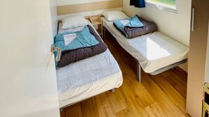 Une petite pièce avec deux lits simples, une étagère en bois au-dessus, une fenêtre avec un rideau et une porte.