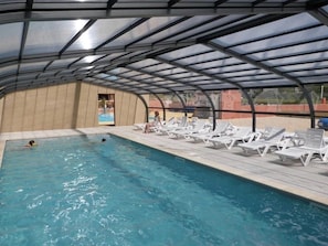 An indoor swimming pool with a covered roof, lounge chairs, and a view of the outside.