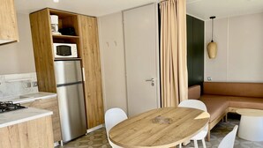 A compact kitchen with a refrigerator, microwave, and stove, a wooden dining table with chairs, a sofa, and a hanging light fixture.