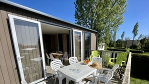 A covered outdoor patio with a table and chairs, a dining area visible through an open door, and a garden with green chairs.