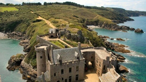 A historic stone castle with a moat, situated on a coastal cliff overlooking the sea.