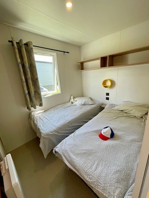 Une petite chambre à coucher, bien rangée, avec deux lits simples, une fenêtre avec des rideaux, une table de chevet et une étagère fixée au mur.
