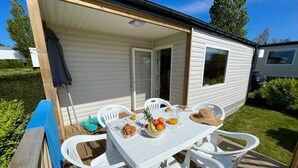 A mobile home with a patio area featuring a table set with breakfast items, white plastic chairs, and a blue mat. - ZEcamping 3* piscine couverte animations locatif TV 28m2 4 pers (Erquy)
