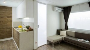 A compact living space with a kitchenette, a sofa, and a window with curtains. - ZEcamping 2* pataugeoire animaux acceptés fitness mobil-home TV clim (Pietra Ligure)