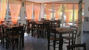 A dining area with tables and chairs, large windows with orange curtains, and a tiled floor. - ZEcamping 2* pataugeoire animaux acceptés fitness mobil-home TV clim (Pietra Ligure)