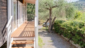 A wooden deck leading to a house with a pergola, surrounded by olive trees and greenery. - ZEcamping 2 étoiles pataugeoire fitness mobil-home TV clim 31m2 6 pers (Pietra Ligure)