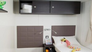 A compact hotel room with a single bed, a bedside table with a lamp, a small shelf with a few items, and a wall-mounted TV. - ZEcamping 2 étoiles pataugeoire fitness mobil-home TV clim 31m2 6 pers (Pietra Ligure)