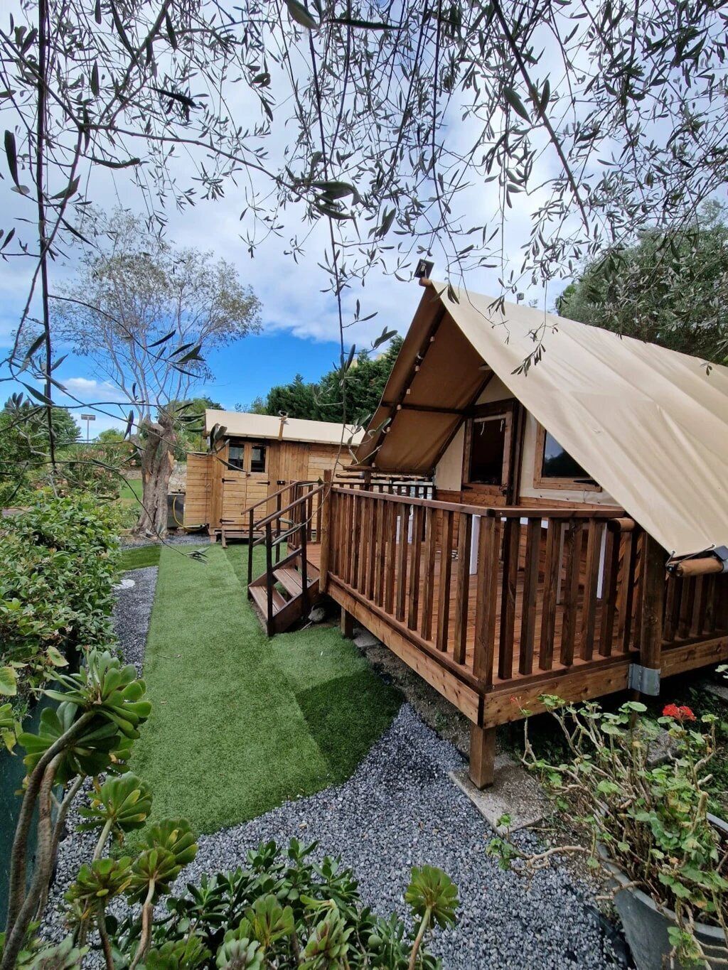 A wooden cabin with a deck, surrounded by greenery and a small lawn.