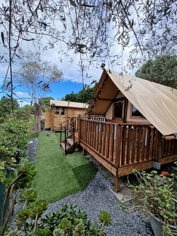 A wooden cabin with a deck, surrounded by greenery and a small lawn.