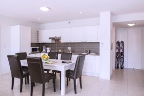 A modern kitchen with white cabinets, a dining table with chairs, and a fruit bowl. - ZEcamping 3 étoiles pataugeoire club enfant locatif TV LV clim 90m2 (Le Sauze-du-Lac)