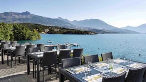 A lakeside restaurant with outdoor seating overlooking a calm lake and mountains. - ZEcamping 3 étoiles pataugeoire club enfant locatif TV LV clim 90m2 (Le Sauze-du-Lac)