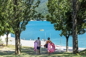 A beach scene with trees, a clear blue sea, and a boat in the distance. - ZEcamping 3 étoiles pataugeoire club enfant locatif TV LV clim 90m2 (Le Sauze-du-Lac)