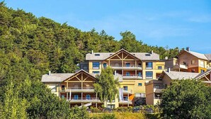A multi-story hotel with balconies, set against a backdrop of dense forest and clear blue sky. - ZEcamping 3 étoiles pataugeoire club enfant locatif TV LV clim 90m2 (Le Sauze-du-Lac)