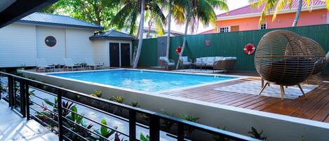 A swimming pool with lounge chairs and a wicker ball chair, surrounded by palm trees and a green fence.