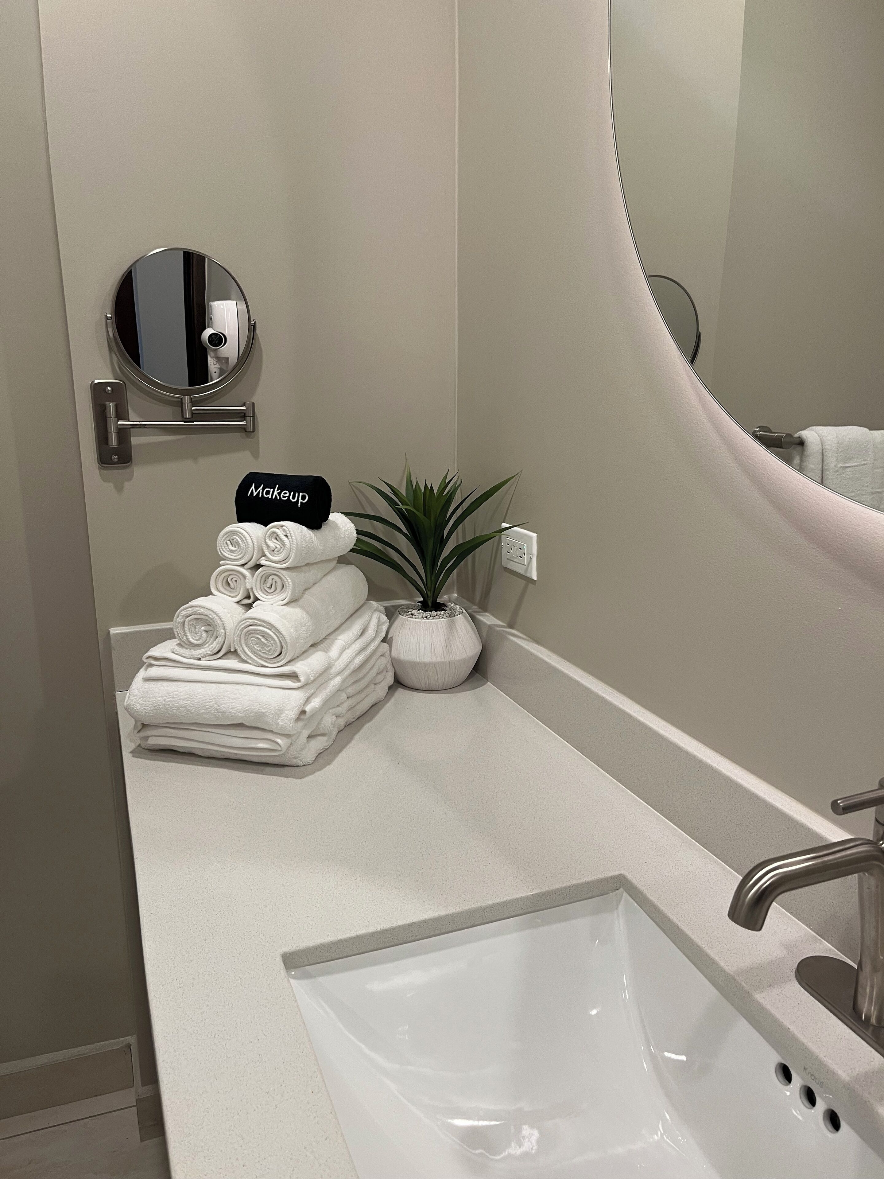 A bathroom with a white sink, a mirror, a towel rack with folded towels, a potted plant, and a sign labeled "Makeup".