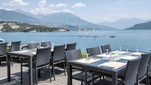 Een terras met rieten tafels en stoelen, met uitzicht op een meer met zeilboten en bergen op de achtergrond.