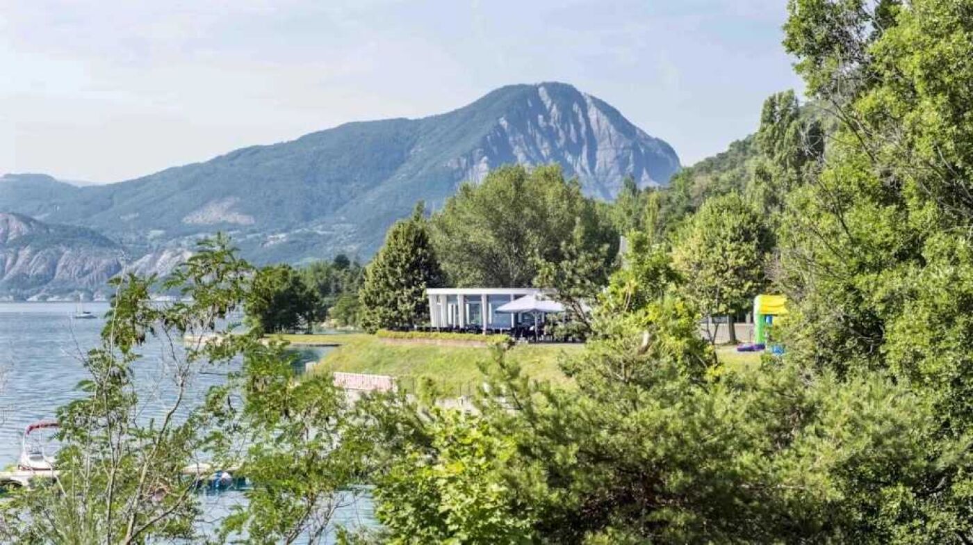 Ein Anwesen am Seeufer mit Bergblick, ein weißes Gebäude mit überdachter Terrasse und ein kleines Gebäude mit gelbem Dach.