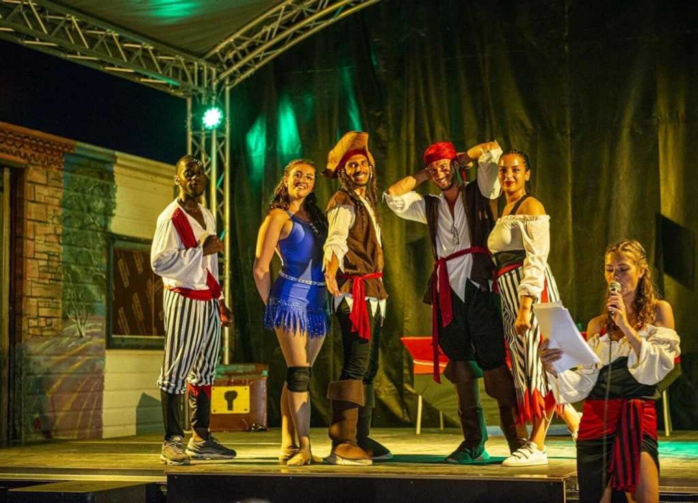 A stage with performers in period costumes, a green curtain backdrop, and a metal truss structure.