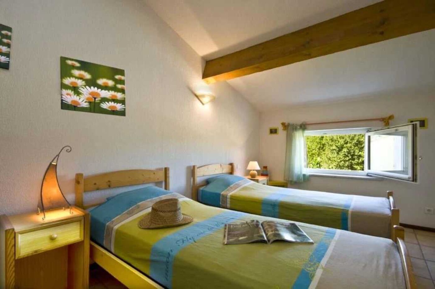 A bedroom with two beds, a window with a view of greenery, a wall-mounted lamp, and a painting of flowers.