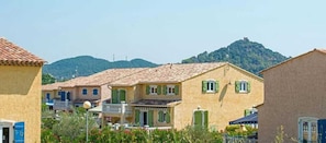 A row of Mediterranean-style buildings with terracotta roofs, green shutters, and a hillside in the background.