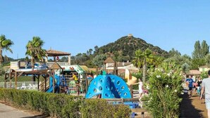 A playground with slides and climbing structures, surrounded by palm trees and a hill in the background.