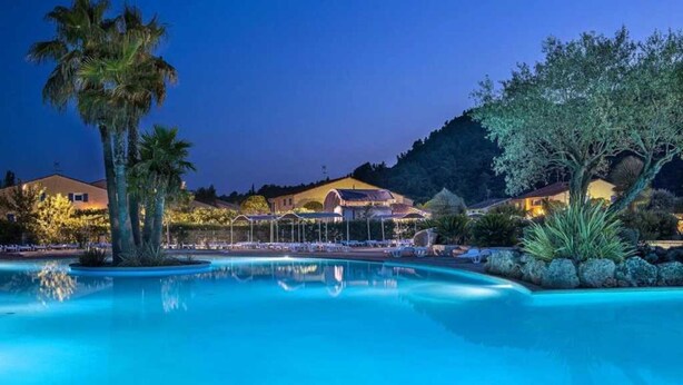 A nighttime view of a resort with a swimming pool, palm trees, and a mountain backdrop.