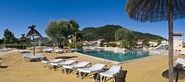 A pool area with sun loungers and umbrellas, surrounded by buildings and trees.