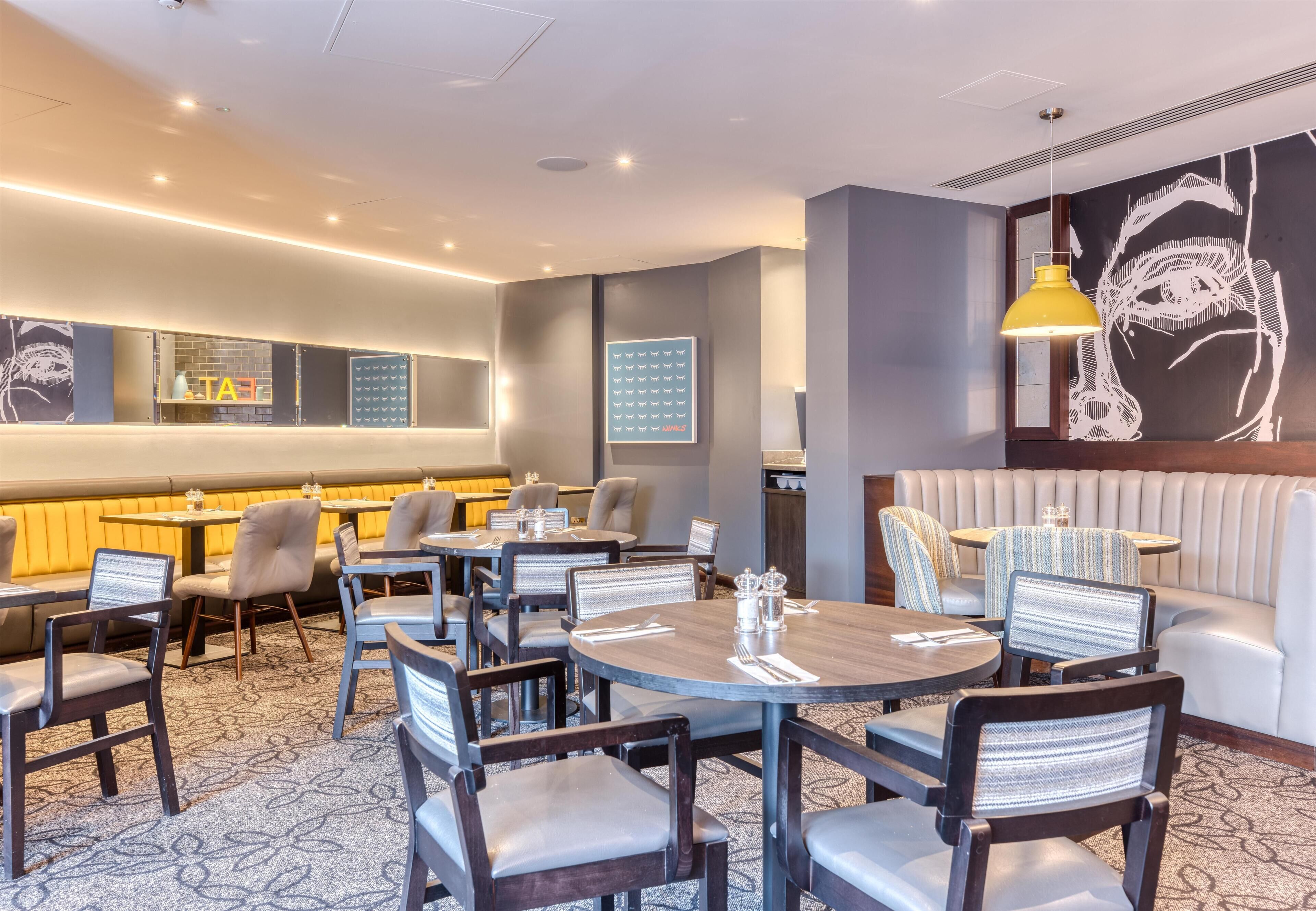 A hotel lobby with round tables, cushioned seating, a large wall art piece, and a modern ceiling design.