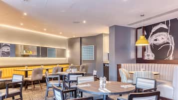A hotel lobby with round tables, cushioned seating, a large wall art piece, and a modern ceiling design.