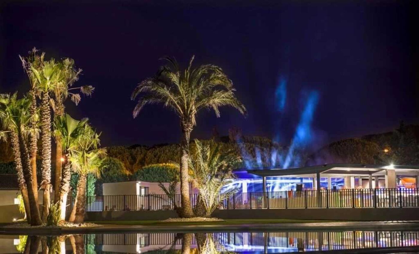 Nachtsicht auf einen Kurort mit Palmen, einem Pool und einem Gebäude mit blau beleuchteten Lichtern.