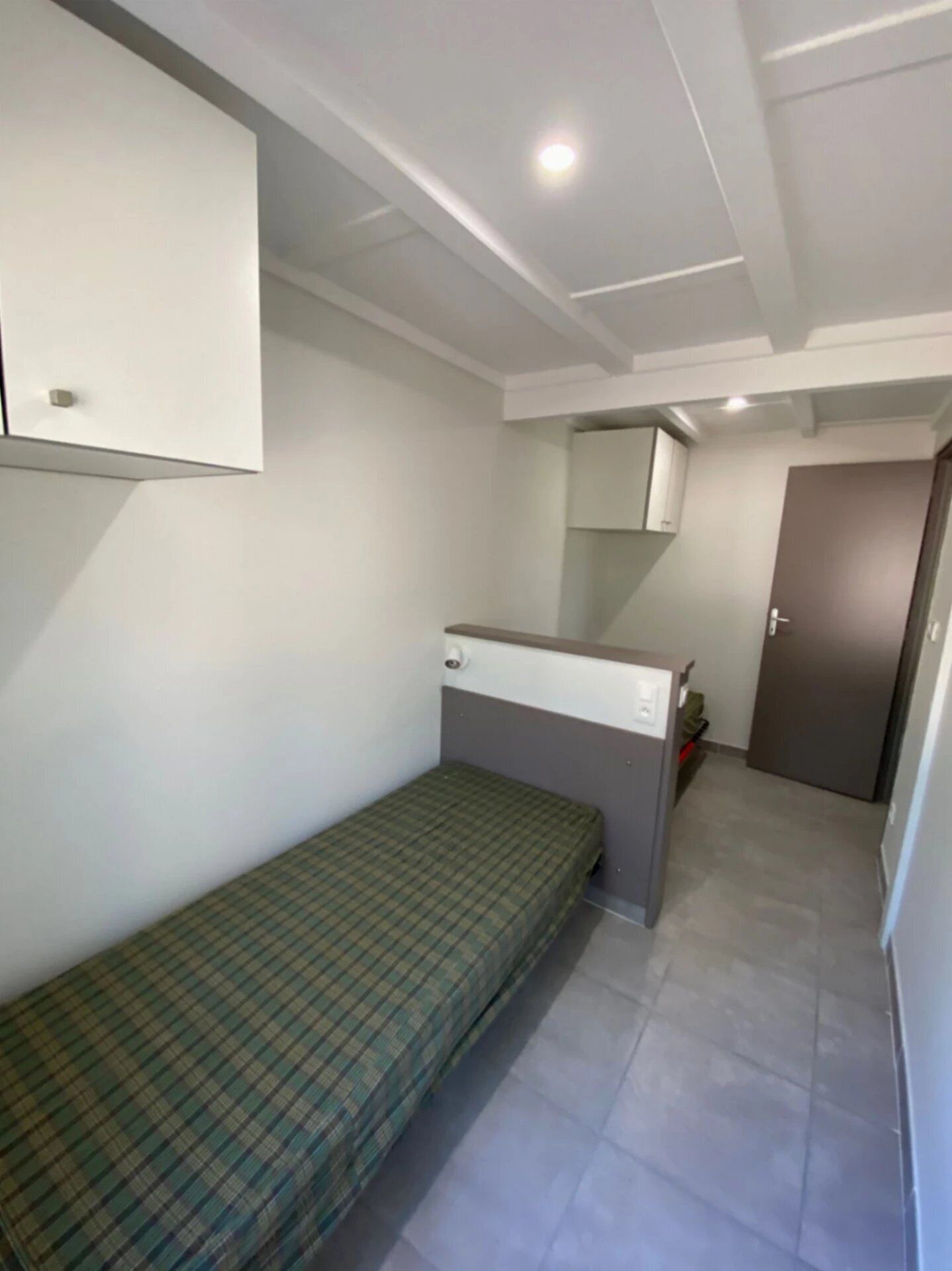 Een kleine eenpersoonskamer met een bed met een groen geruit sprei, een witte kast, een klein bureau en een deur.