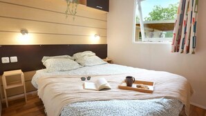 A neatly made bed with a book, sunglasses, and a mug on a tray, a small bedside table, and a window with curtains. - ZEcamping 3* piscine animaux acceptés fitness locatif 45m2 6 pers (Vensac)
