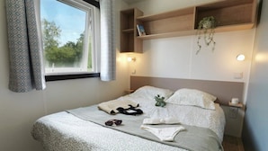 A neatly made bed with a patterned bedspread, a pair of sunglasses, and a folded towel on the bed. There is a window with patterned curtains, a bedside table with a lamp, and a shelf above the bed with a plant.
