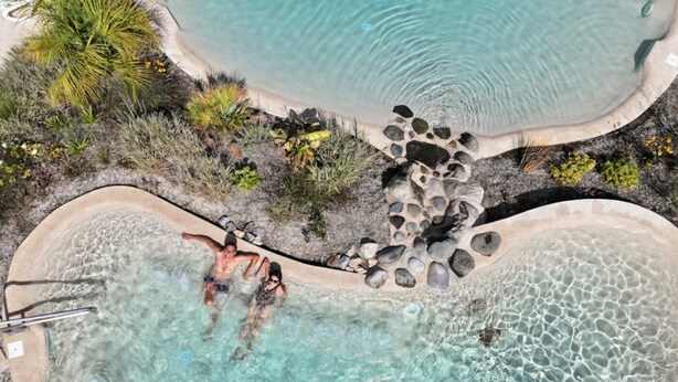An aerial view of a pool area with clear water, surrounded by rocks and tropical vegetation.
