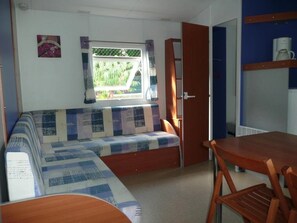 A compact RV interior with a sofa bed, a dining table, and a window with a view of greenery.