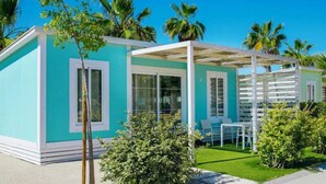 A modern, two-story building with a teal exterior and white trim. It features large windows, a covered patio area with seating, and is surrounded by lush greenery and palm trees. - ZEcamping 5* familial espace aquatique locatif TV LV clim 32m2 5 pers (Sant Felip Neri)