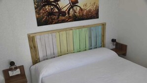 A bed with a wooden headboard painted in multiple colors, a white bedspread, and two bedside tables with lamps. - ZEcamping 5* familial espace aquatique locatif TV LV clim 32m2 5 pers (Sant Felip Neri)