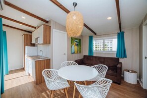 A compact living space with a kitchenette, a dining area with white chairs, a brown sofa, and a large woven light fixture.